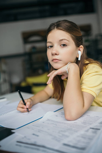 nastolatka dziewczyna w bezprzewodowy słuchawki trzymając długopis i patrząc w aparat podczas odrabiania lekcji - Zdjęcie, obraz