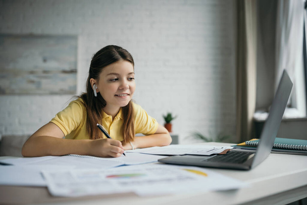 χαμογελαστό κορίτσι σε ασύρματο ακουστικό κρατώντας στυλό και κοιτάζοντας το φορητό υπολογιστή στο σπίτι - Φωτογραφία, εικόνα