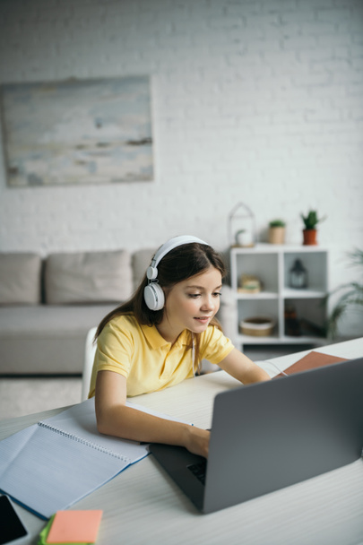θετική μαθήτρια σε ακουστικά χρησιμοποιώντας φορητό υπολογιστή στο σαλόνι στο σπίτι - Φωτογραφία, εικόνα