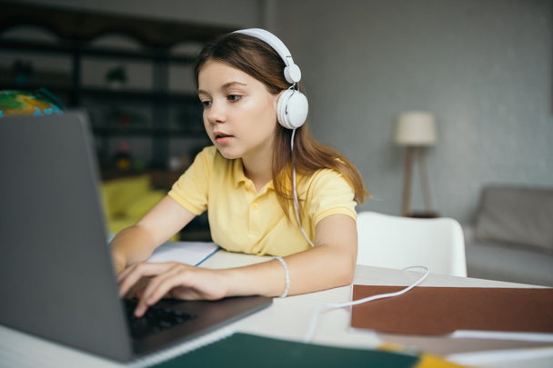 Brünette Schülerin mit kabelgebundenen Kopfhörern lernt zu Hause und tippt auf Laptop  - Foto, Bild