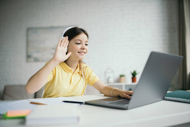χαμογελαστό κορίτσι με ακουστικά κουνώντας το χέρι κοντά στο laptop κατά τη διάρκεια online μαθήματος στο σπίτι - Φωτογραφία, εικόνα
