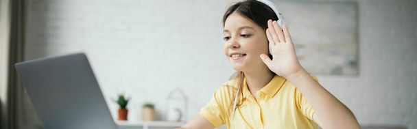 девочка-подросток в наушниках машет рукой и улыбается во время видеозвонка на ноутбуке, баннер - Фото, изображение