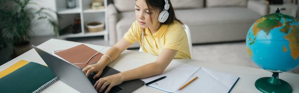 προβολή υψηλής γωνίας του κοριτσιού σε ακουστικά δακτυλογράφησης σε φορητό υπολογιστή κοντά σε φορητούς υπολογιστές και υδρόγειο, banner - Φωτογραφία, εικόνα