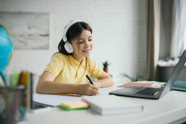 smiling schoolgirl writing in notebook while sitting in headphones near laptop at home - Photo, Image