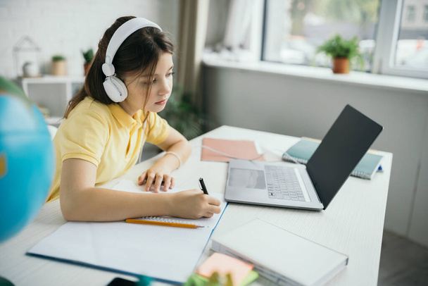Kulaklıklı çocuk dizüstü bilgisayara bakıyor ve evde deftere yazıyor. - Fotoğraf, Görsel