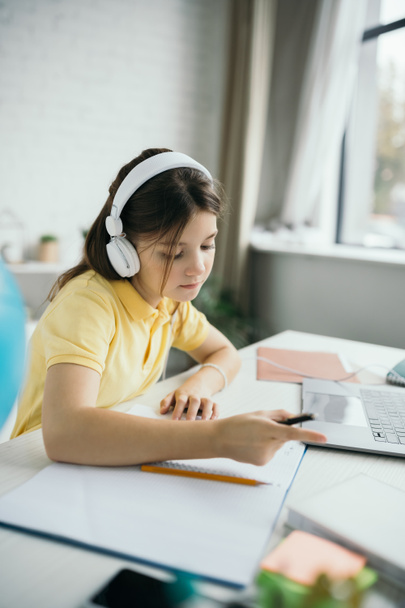 schoolgirl in headphones pointing with finger while studying near blurred laptop at home - Фото, изображение