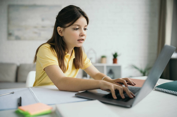 preteen girl typing on laptop near blurred copybooks during homeschooling - Zdjęcie, obraz
