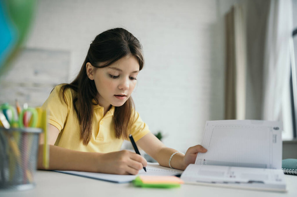 niña preadolescente escribiendo en cuaderno cerca del diario en primer plano borroso - Foto, Imagen