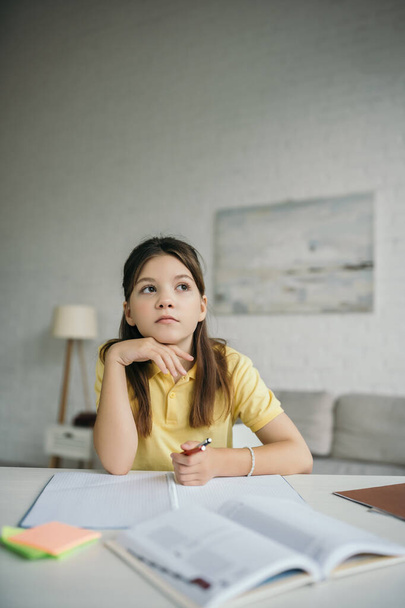 pozorná dívka drží ruku u brady a dívá se stranou, zatímco dělá domácí úkol - Fotografie, Obrázek
