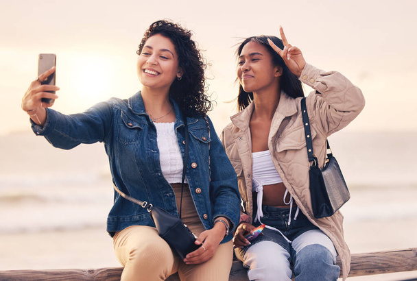 Selfie, peace sign and women with phone, beach and relax outdoor for fun, travel and smile together. Female friends, young girls and sunset with smartphone, hand gesture and social media to connect - Фото, изображение