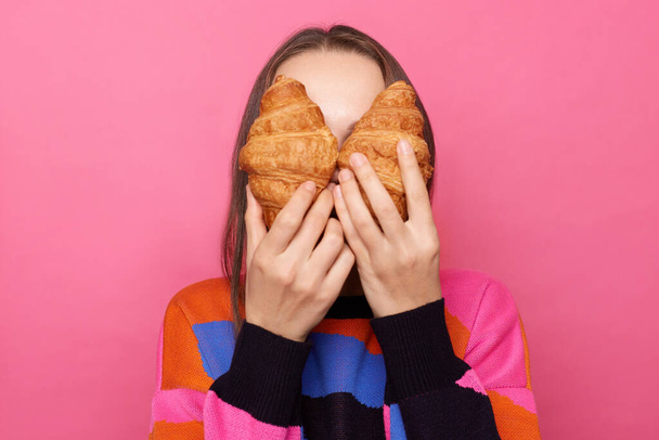 Portrait of unknown woman with dark straight hair wearing jumper standing isolated over pink background, covering her eyes with croissants, hiding her face. - Foto, Imagen
