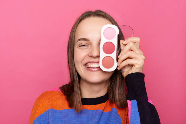 Image of joyful cheerful woman with brown hair wearing sweater, covering her eye with eyeshadow palette, looking at camera with toothy smile, posing isolated over pink background. - Photo, Image