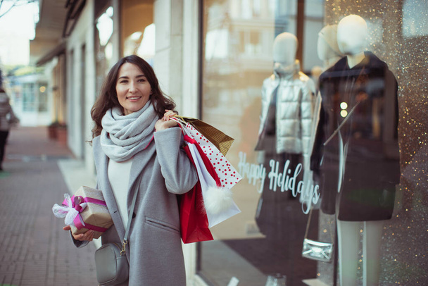 Young happy smiling woman holding shopping bags and Christmas presents in her hands. Concept of European winter holidays, sales and gifts, or black friday. - Foto, afbeelding