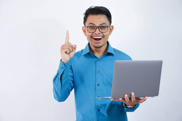 ásia homem com óculos saiu ou feliz obter novo ideias apontando dedo para cima e segurando laptop vestindo azul camisa isolado no branco fundo - Foto, Imagem