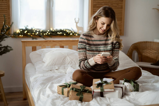 Blanke vrouw met behulp van telefoon tijdens het zitten op bed tussen kerstcadeaus - Foto, afbeelding