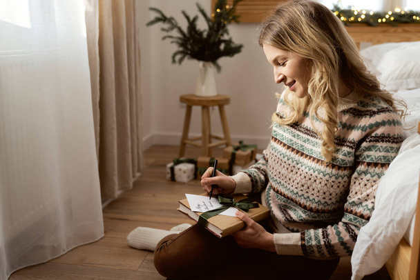 Fröhliche Kaukasierin sitzt auf dem Fußboden und schreibt Weihnachtskarte zwischen Geschenken - Foto, Bild