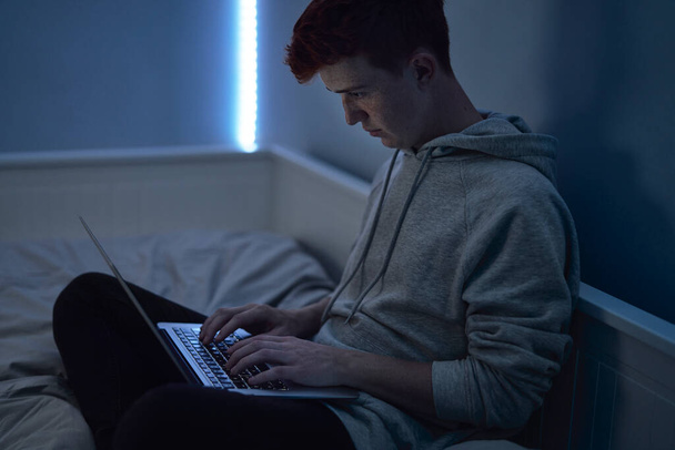 Enfoque caucásico adolescente usando el ordenador portátil mientras está sentado por la noche en su habitación  - Foto, Imagen