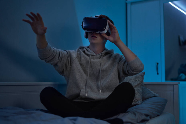 tiener Kaukasische jongen met behulp van virtual reality bril terwijl zitten in de slaapkamer 's nachts - Foto, afbeelding