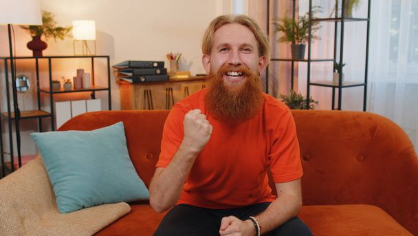 Happy bearded redhead man shouting, celebrating success, winning, goal achievement, good news, raising fists in gesture I did it, lottery luck at home apartment. Young guy indoors in room on couch - Photo, Image