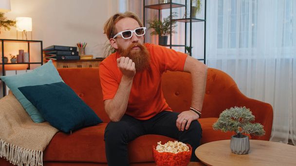 Portrait of bearded excited young man sits on sofa eating popcorn and watching interesting tv serial, sport game, film, online social media movie content at home. Guy enjoying domestic entertainment - Fotó, kép