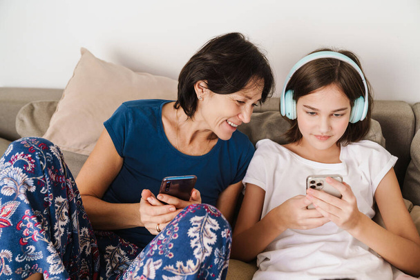 White woman and her daughter using gadgets while resting on couch at home - Foto, Imagem