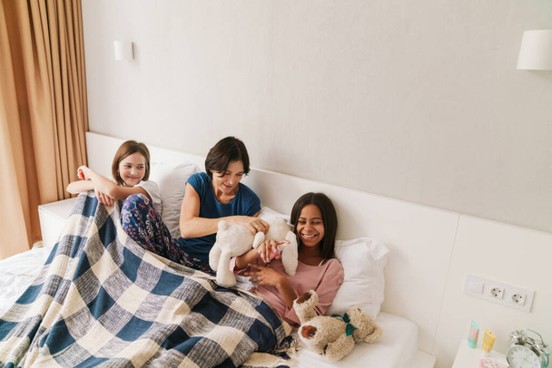 Femme blanche et ses filles se moquent des jouets tout en se reposant sur le lit à la maison - Photo, image