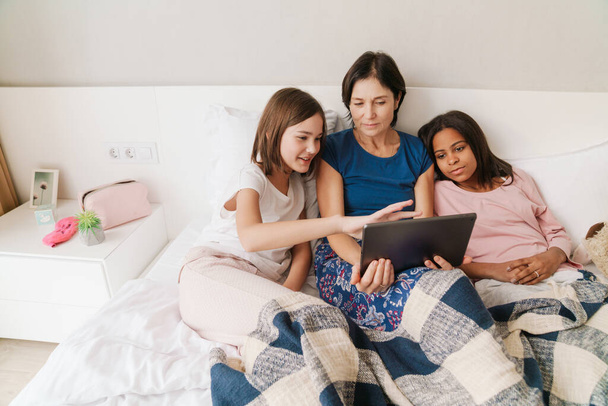 White woman and her daughters using tablet computer while resting on bed at home - 写真・画像