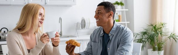 complacida pareja multiétnica con taza de café y delicioso croissant mirándose en la cocina, pancarta - Foto, imagen