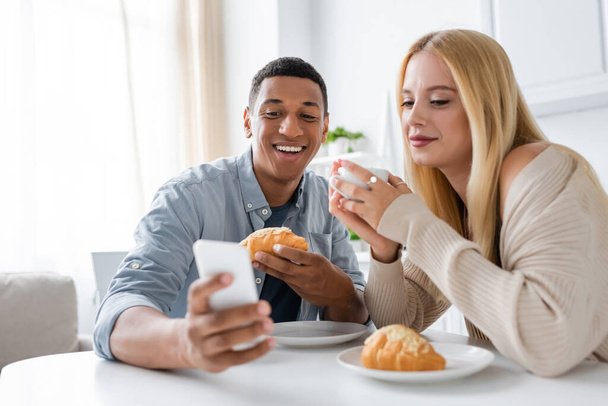 cheerful african american man showing blurred smartphone to smiling girlfriend during breakfast in kitchen - 写真・画像