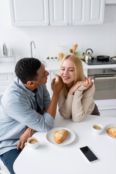 african american man touching face of pretty blonde girlfriend near coffee cups and croissants in kitchen - Фото, изображение