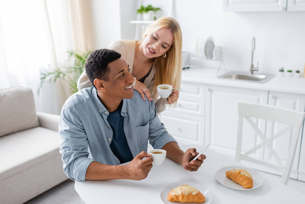 blonde woman holding coffee cup near african american man using smartphone during breakfast in kitchen - Fotografie, Obrázek