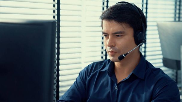 Young competent asian male call center agent working at his computer while simultaneously speaking with customers. Concept of an operator, customer service agent working in the office with headset. - Photo, image