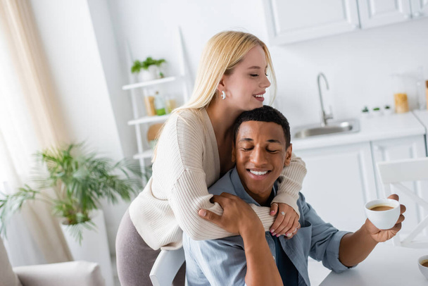 happy blonde woman embracing african american boyfriend smiling with closed eyes and holding coffee cup - Fotoğraf, Görsel