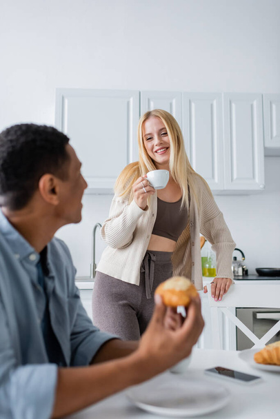 pretty blonde woman with coffee cup looking at blurred african american boyfriend having breakfast in kitchen - Foto, afbeelding