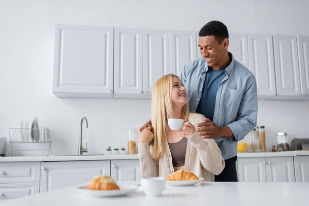 african american man smiling at young blonde girlfriend sitting with coffee cup near blurred croissants - Photo, Image