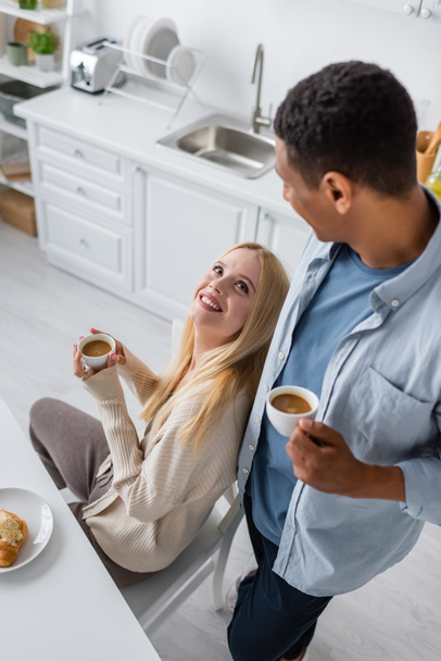 high angle view of blonde woman looking at african american boyfriend while drinking morning coffee in kitchen - Φωτογραφία, εικόνα