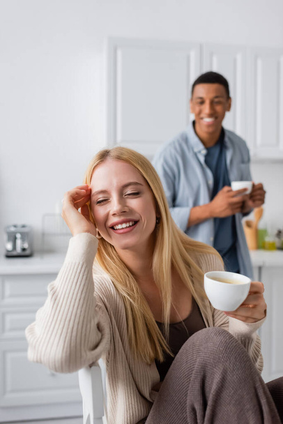 young and happy woman with closed eyes holding cup of coffee near african american boyfriend smiling on blurred background - Foto, Imagem
