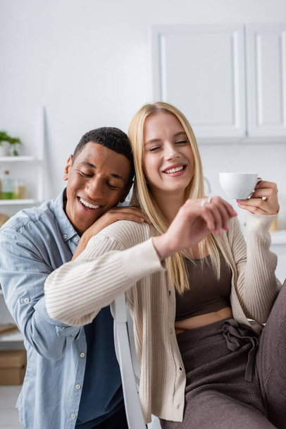 pleased african american man with closed eyes embracing blonde girlfriend holding coffee cup  - 写真・画像