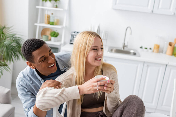 african american man smiling near young blonde girlfriend sitting with morning coffee in kitchen - Foto, afbeelding