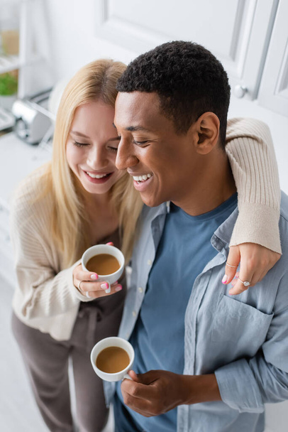 Kapalı gözlü sarışın kadın ve mutfakta kahve fincanlarıyla dikilen mutlu Afro-Amerikan adam. - Fotoğraf, Görsel