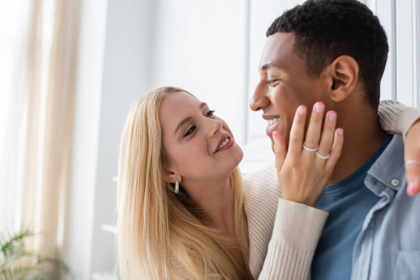 happy blonde woman hugging african american boyfriend and touching his face in kitchen - Foto, Imagem
