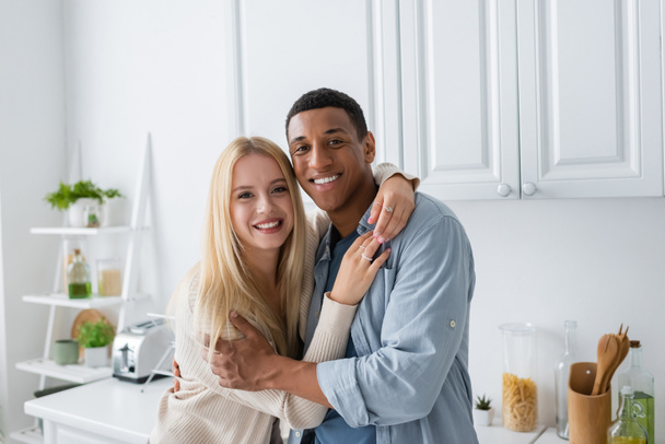 cheerful interracial couple looking at camera while embracing in kitchen - Photo, Image