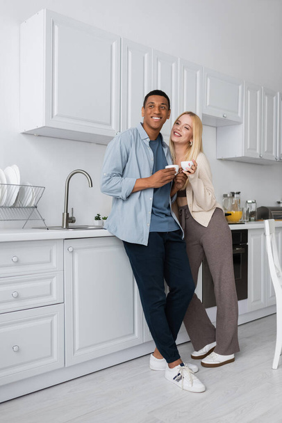 full length of happy interracial couple standing with coffee cups in modern kitchen - Zdjęcie, obraz