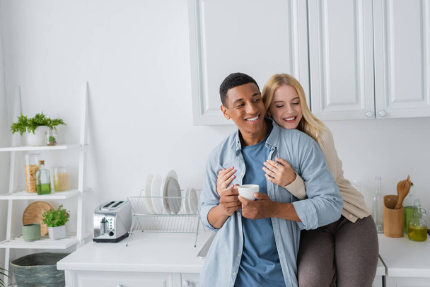 joyful blonde woman with closed eyes sitting on kitchen worktop and hugging african american boyfriend standing with coffee cup - Photo, Image