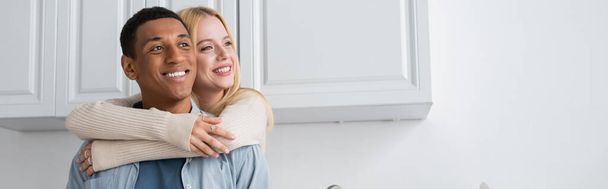 young blonde woman hugging cheerful african american boyfriend and looking away in kitchen, banner - Foto, Imagem