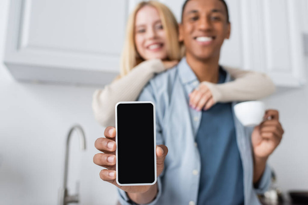 enfoque selectivo de teléfono inteligente con pantalla en blanco en la mano del hombre afroamericano borroso cerca de la mujer sonriente - Foto, Imagen