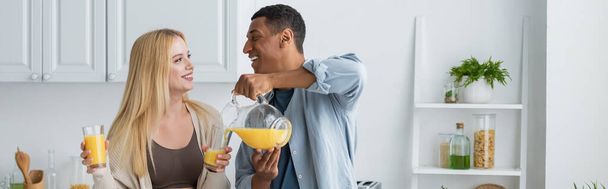 陽気なアフリカ系アメリカ人男性見ブロンドガールフレンドと注ぎます新鮮なオレンジジュースでキッチン,バナー - 写真・画像