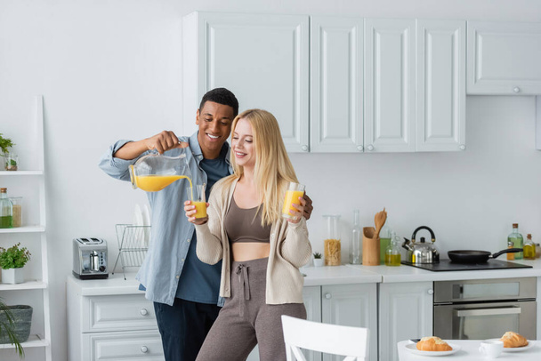 gelukkig Afrikaans amerikaanse man gieten sinaasappelsap in de buurt mooi vriendin holding bril in keuken - Foto, afbeelding