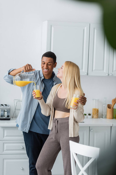 cheerful african american man pouring orange juice near young blonde girlfriend standing with glasses in kitchen - Фото, зображення