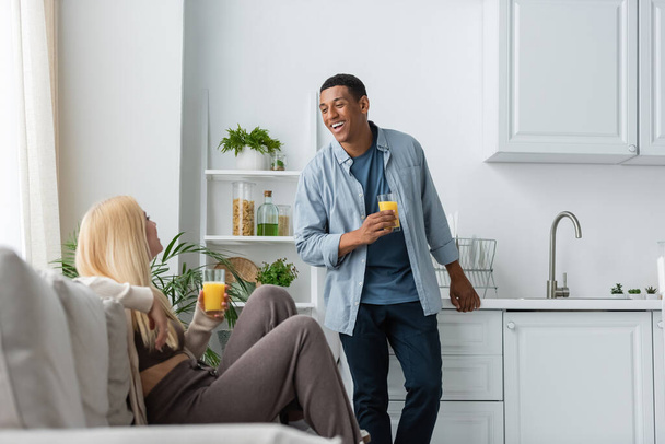 happy african american man with glass of fresh orange juice standing near girlfriend sitting on blurred couch in kitchen - Foto, Imagen